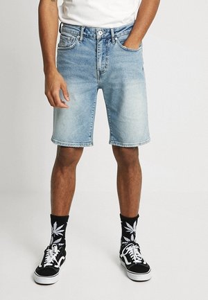 Homme portant un short en jean bleu clair, des chaussettes noires avec des motifs de feuilles blanches, des baskets noires et blanches, et une chemise blanche avec une main dans la poche.