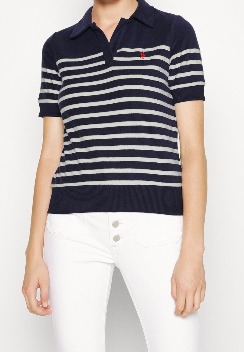 Femme portant un polo à manches courtes rayé bleu marine et blanc avec un pantalon blanc et deux boutons visibles.