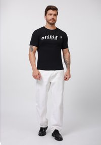 Black short-sleeve t-shirt featuring a white evolution graphic. Paired with white trousers and black shoes, showing a relaxed fit.