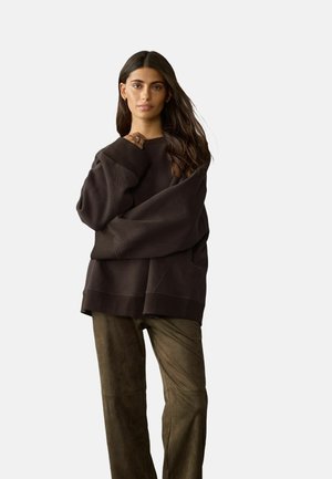 Woman with long dark hair wearing a loose dark brown sweatshirt and olive green pants against a plain light background.