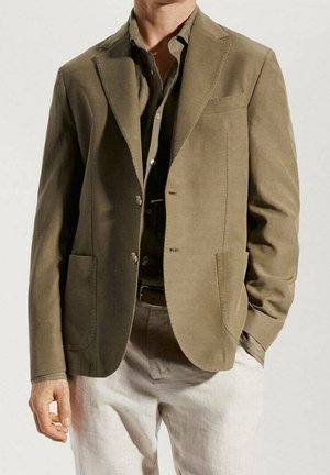 Hombre con blazer color verde oliva sobre camisa a juego y pantalones beige, con una mano en el bolsillo y la otra relajada al costado.