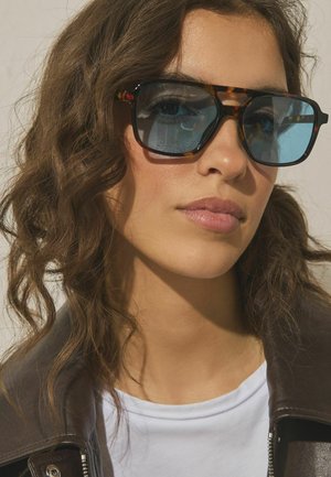 Prim-plan cu o femeie cu păr creț, purtând ochelari de soare cu ramă de țestoasă și lentile albastre, o cămașă albă și o jachetă întunecată din piele.