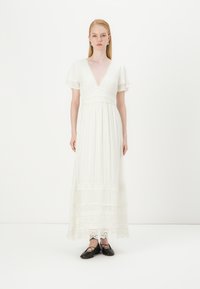 Vestido maxi branco com mangas curtas e largas, decote V profundo, cintura franzida e detalhes de renda na barra; combinado com sapatos pretos rasos.