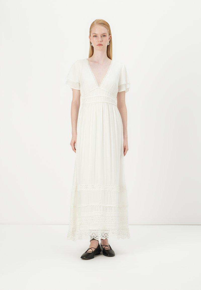 Vestido maxi branco com mangas curtas e largas, decote V profundo, cintura franzida e detalhes de renda na barra; combinado com sapatos pretos rasos.