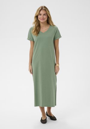 Femme aux cheveux blonds longs portant une robe maxi à manches courtes vert sauge et des ballerines noires tissées, debout devant un fond blanc uni.