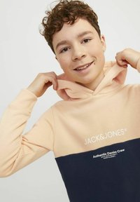 Mikina s kapucňou v broskyňovej a námorníckej modrej farbe, s mäkkou textúrou, kontrastnými farbami a s logom na hrudi.