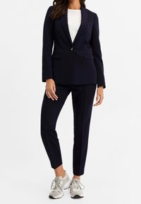 Marinblå skräddarsydd kostym med enbladsblazer med slag och matchande slim-fit byxor. Kombinationen med ljusgrå sneakers.