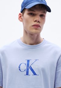 Jasnoniebieska bawełniana koszulka z dużym, niebieskim logo "CK" i białym napisem "Calvin Klein", uzupełniona o okrągły dekolt.