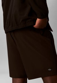 Personne portant un short noir et une veste assortie, une main dans la poche, montrant un logo Calvin Klein sur le short.