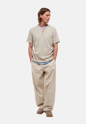 Homme aux cheveux longs portant un t-shirt henley beige à manches courtes, un sous-vêtement bleu clair, un pantalon beige ample et des chaussures marron, debout les mains dans les poches.