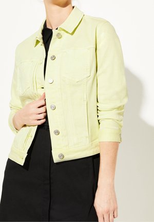 Personne portant une veste boutonnée jaune clair sur une robe noire, une main ajustant la veste près de la poitrine.