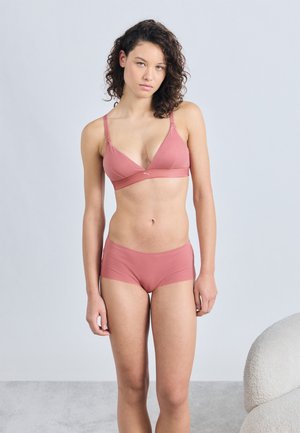 Νεαρή γυναίκα με σγουρά μαλλιά στέκεται φορώντας ταιριαστό στητό dusty pink σουτιέν και σορτς μπροστά σε απλό φωτεινό φόντο.