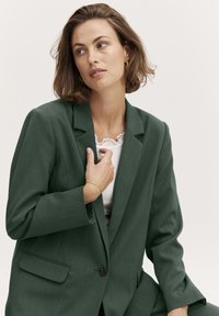 Zaļš strukturēts žakets ar izgriezumu pie apkakles, pogu aizdari un divām priekšējām kabatām. Vilkts pār krēmkrāsas augšdaļu ar mežģīņu apdari un apvienots ar delikātu rokassprādzi.