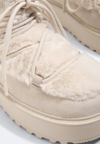 Botte d'hiver beige avec semelle plateforme épaisse, dessus doux et duveteux, accents en daim et lacets ronds beige noués en boucles.