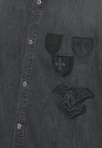 Σκούρο τζιν πουκάμισο με ασημένια κουμπιά, που φέρει τρία κεντημένα μαύρα μπαλώματα: δύο σχέδια με ασπίδες και ένα έμβλημα αετού.