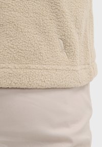 Giacca in pile color crema con una superficie morbida e texture. Presenta un discreto dettaglio del logo vicino all'orlo. Abbinata a un tessuto beige chiaro sotto.