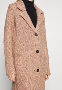 Abrigo de punto en tono melocotón con acabado texturizado, que presenta un cuello con muesca, tres botones negros y dos bolsillos frontales.