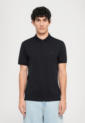 Young man with curly hair wears a fitted black polo shirt and light blue jeans, standing against a plain white background.