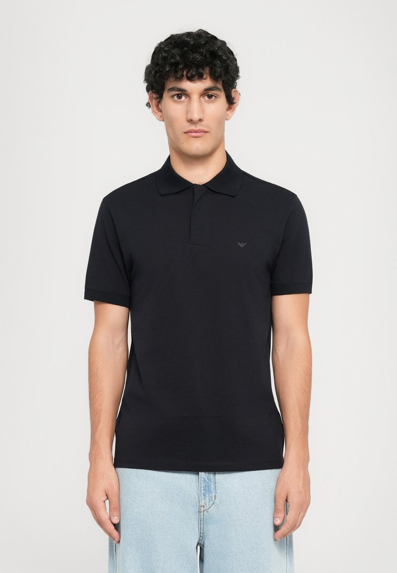 Ung man med lockigt hår bär en tät svart polotröja och ljusblå jeans, stående mot en enfärgad vit bakgrund.