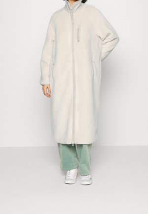 Persoon draagt een knielange, lichtbeige fleecejas met rits, muntgroene broek en witte sneakers met roze accenten, staand tegen een effen achtergrond.