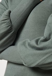 Zielony prążkowany sweter z bufkami na rękawach. Materiał wydaje się miękki z subtelnym połyskiem. Zbliżenie na detale rękawa i fakturę.