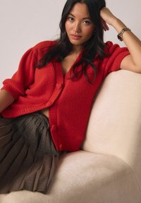 Rdeč pleten kardigan s kratkimi rokavi in sprednjimi gumbi, kombiniran z rjavo karo krilo z gubami. Sedi na mehki bež podlagi.