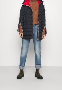 Osoba ubrana w czarną pikowaną kurtkę z czerwonym podbiciem, brązowy sweter dziany, podwinięte niebieskie dżinsy i ciemne masywne botki, stojąca na tle bez wzorów.