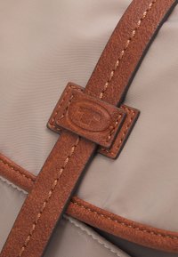 Ruskea nahkahihna ommelluilla yksityiskohdilla pitää kiinni vaaleanbeigestä kankaasta, jossa on logo suorakaiteen muotoisessa nahkapalassa.