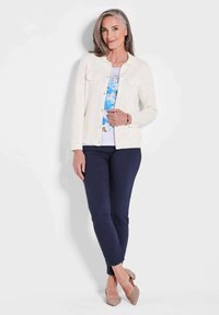 Witte jas over een top met bloemenprint, marineblauwe broek en beige hakken. Model heeft lang grijs haar en staat tegen een eenvoudige achtergrond.