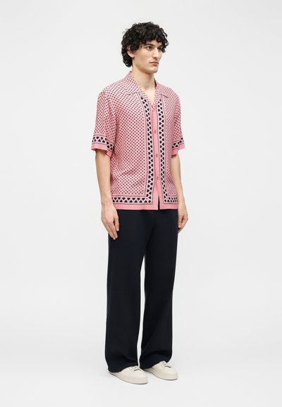 Νέος άνδρας με σγουρά μαλλιά, που φοράει ροζ πουκάμισο με κουμπιά και μαύρο σχέδιο, μαύρο φαρδύ παντελόνι και λευκά αθλητικά παπούτσια, στέκεται ακίνητος.