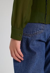 Blusa verde oliva con una texture trasparente, dotata di polsini con bottoni. Indossata con jeans in denim blu dal taglio classico e con tasche posteriori.