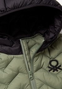 Giacca imbottita con corpo verde e cappuccio nero, chiusura con zip, texture trapuntata e logo ricamato nero sul petto.