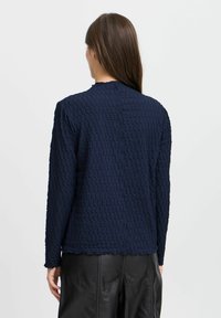 Haut à manches longues texturé bleu marine avec un motif géométrique, doté d'un col rond et d'un ourlet droit, porté avec un pantalon en cuir noir.