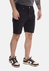 Shorts in denim nero con una vestibilità regolare, cinque tasche e dettagli di cucitura sottili, abbinati a sneaker marroni e crema.