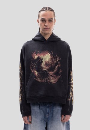 Person med fletninger iført en sort hættetrøje med fantasystil grafik og tekst, stående mod en ensfarvet grå baggrund.