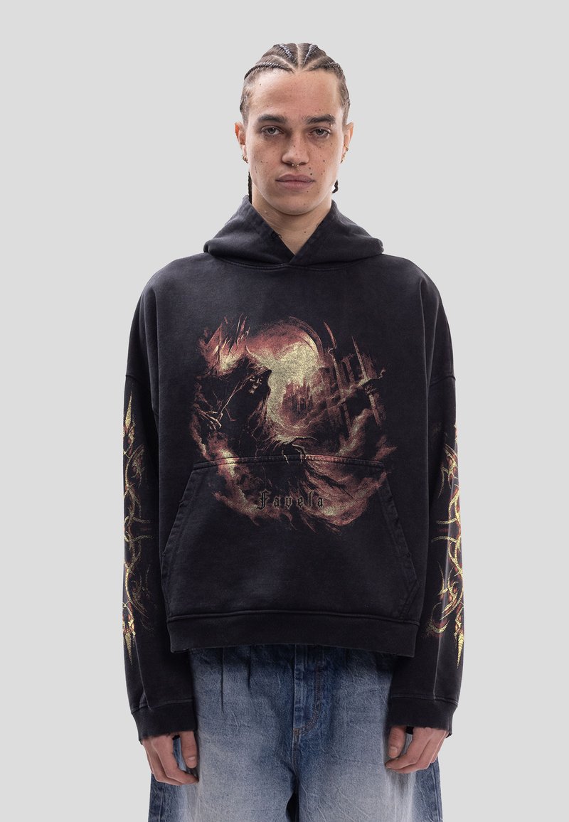 Personne aux cheveux tressés porte un sweat à capuche noir avec un graphisme de style fantastique et du texte, se tenant devant un fond gris uni.