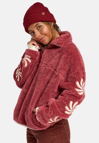 Giacca in pile color rosa polveroso con colletto alto e dettagli ricamati bianchi sulle maniche. Il modello indossa un berretto di maglia rosso.