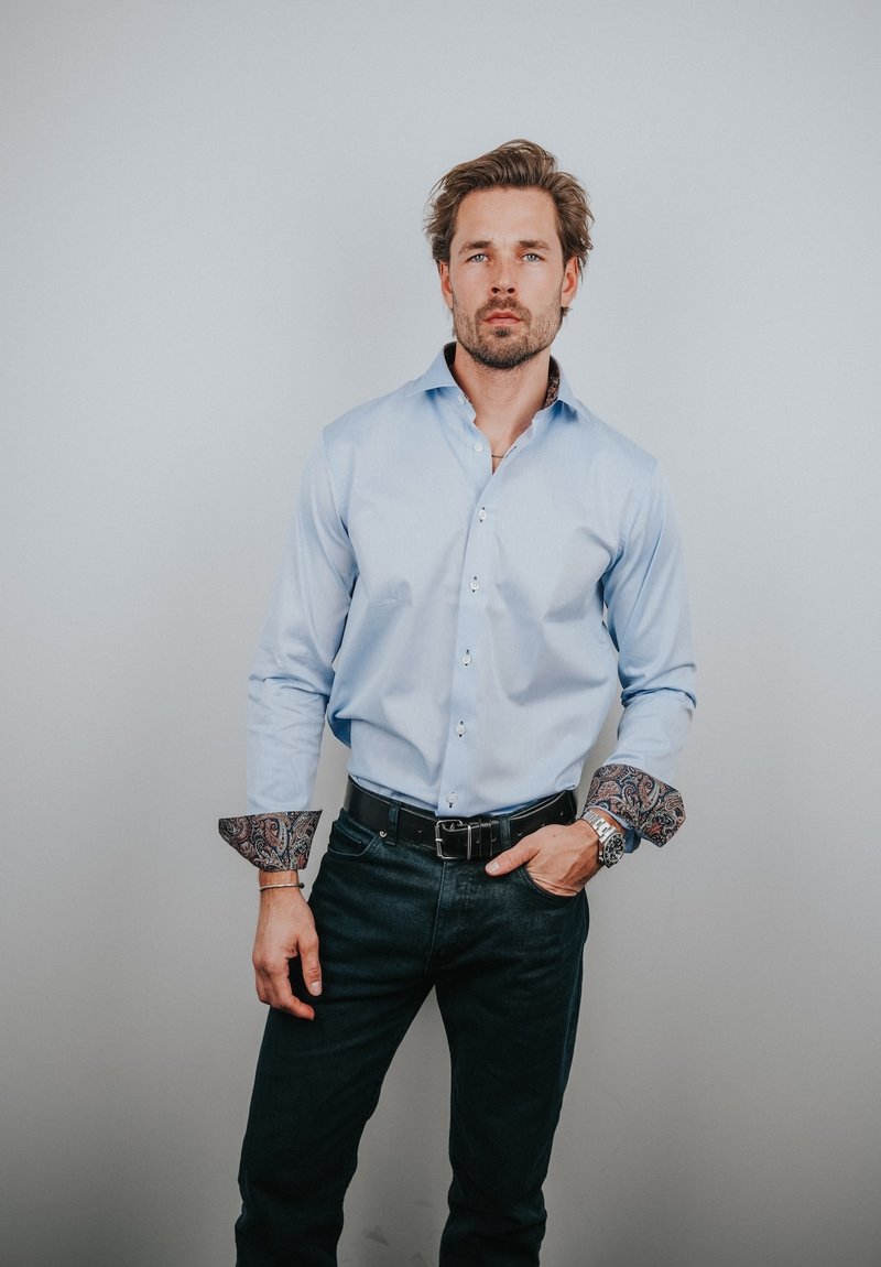 Chemise bleu clair avec poignets et col foncés à motifs, portée sur un jean foncé et maintenue par une ceinture noire. Présente des boutons sur le devant.