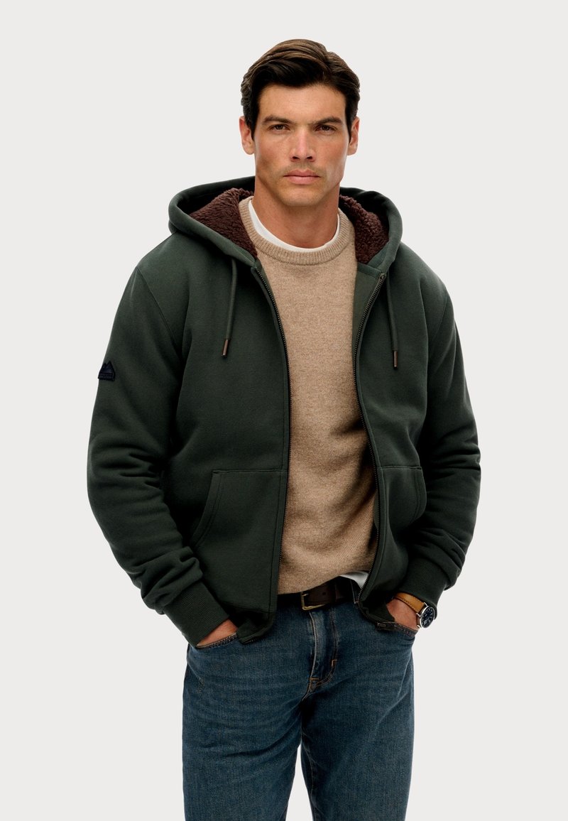 Groene zip-up hoodie met een bruine fleece voering, gedragen over een beige sweater. Voorzien van trekkoorden en voorzakken, gecombineerd met blauw denim.