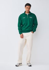 Grøn sweatshirt med kvart-lynlås design, ribstrikkede manchetter og broderet tekst foran, kombineret med off-white bukser og grå sneakers.
