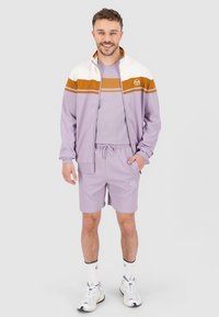 Mees, kes kannab lavendlit ja pruuni spordijopet, sobivaid lühikesi pükse, valgeid sokke ja valgeid tossusid, seisab ja naeratab.