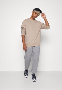 Beżowa bluza z długimi rękawami, logo na klatce piersiowej, szare dresowe spodnie o tapered kroju oraz czapka w kolorze czarnym. Czarno-białe sneakersy z niebieskimi akcentami.