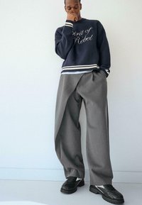 Marineblauwe trui met de tekst "Spirit of Rebel", witte strepen bij de manchetten en zoom, gecombineerd met losse grijze broek en zwarte sneakers.