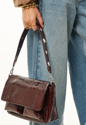 Mano sosteniendo un bolso marrón oscuro, brillante, con textura de reptil y tachuelas de metal, llevado con jeans de mezclilla azul claro y pulseras doradas en la muñeca.