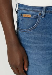 Los jeans de mezclilla azul Wrangler presentan un ajuste clásico, costuras de cobre, parche de cuero con el logo y un primer plano de una mano reposando sobre la tela.