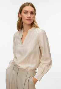 Krēmveida blūze no viegla auduma ar V veida kakla izgriezumu, gariem piedurknēm un smalkām vertikālām svītrām. Kombinēta ar gaiši bēšām biksēm.