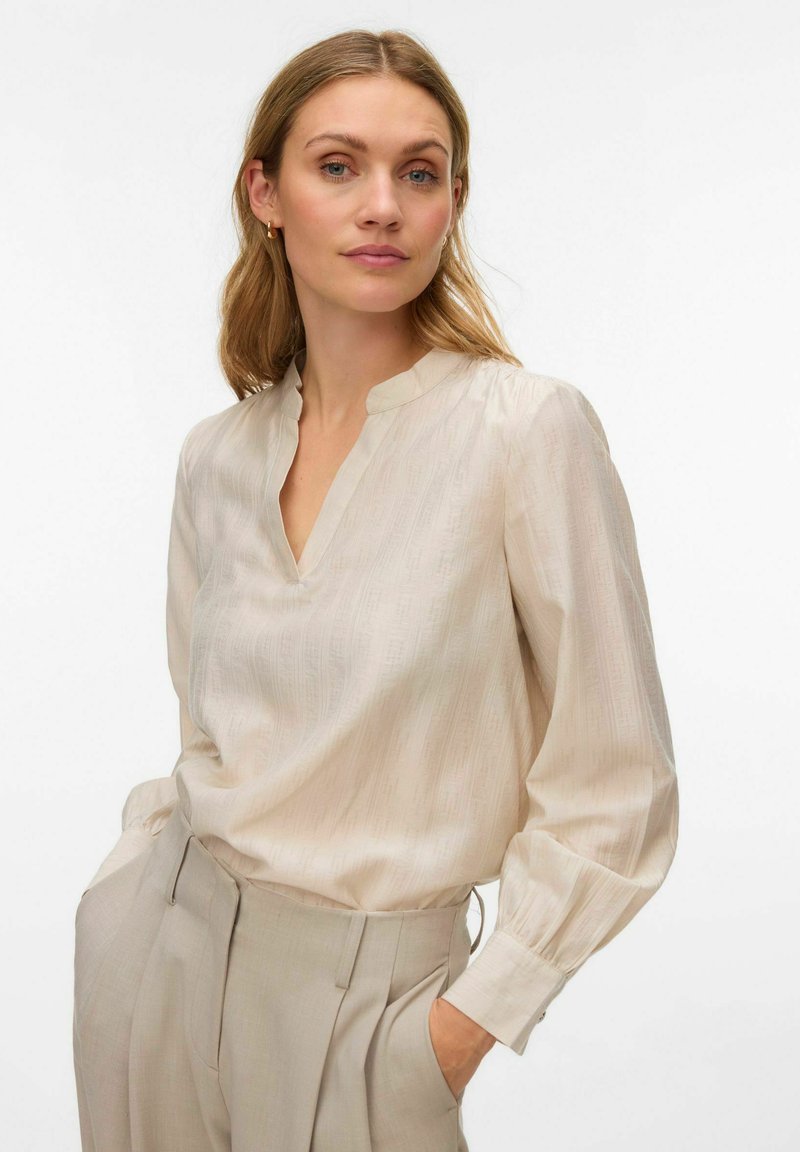 Krēmveida blūze no viegla auduma ar V veida kakla izgriezumu, gariem piedurknēm un smalkām vertikālām svītrām. Kombinēta ar gaiši bēšām biksēm.