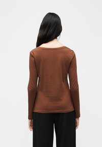 Γυναίκα με μακριά μαύρα μαλλιά που φοράει καφέ μπλούζα με μακριά μανίκια και μαύρο παντελόνι, γυρισμένη προς τα πίσω με φόντο ένα απλό φωτεινό φόντο.