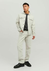 Giubbotto in denim beige chiaro sopra una camicia grigia scura, abbinato a jeans chiari e sneaker nere. Presenta due tasche sul petto e chiusura a bottoni.