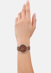 Brauner Lederarmbanduhr mit einem Holz-Zifferblatt, silberner Edelstahl-Lünette und minimalistischen Stundenmarkierungen. Hand gezeigt, die die Uhr trägt.